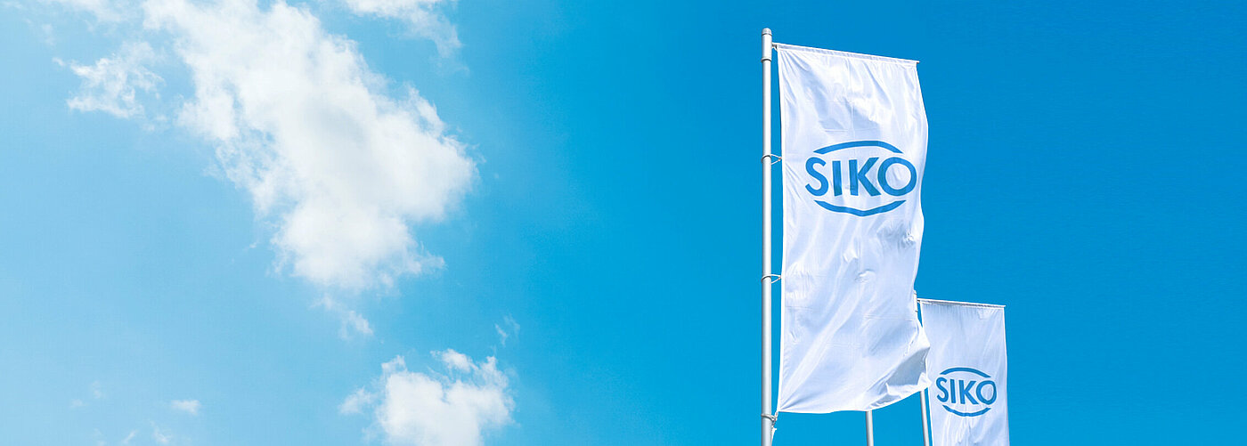 Weiße Siko-Flagge mit blauem Logo vor blauem Himmel mit Sonnenschein